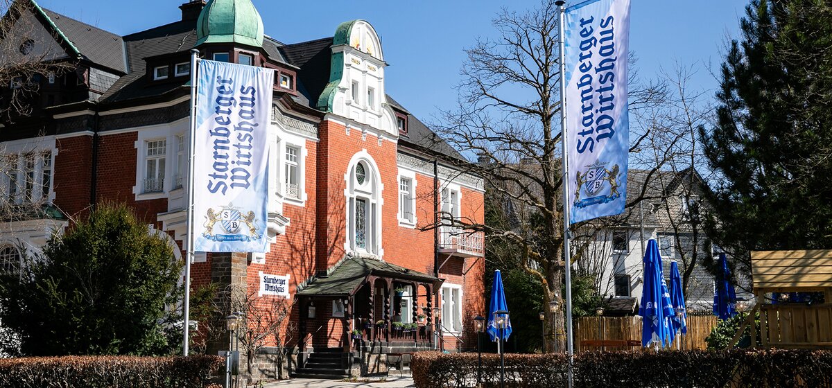 Außenansicht des historischen Gebäudes des Starnberger Wirtshauses in Olpe mit zwei Fahnen und Außenbereich mit Tischen und blauen Sonnenschirmen.