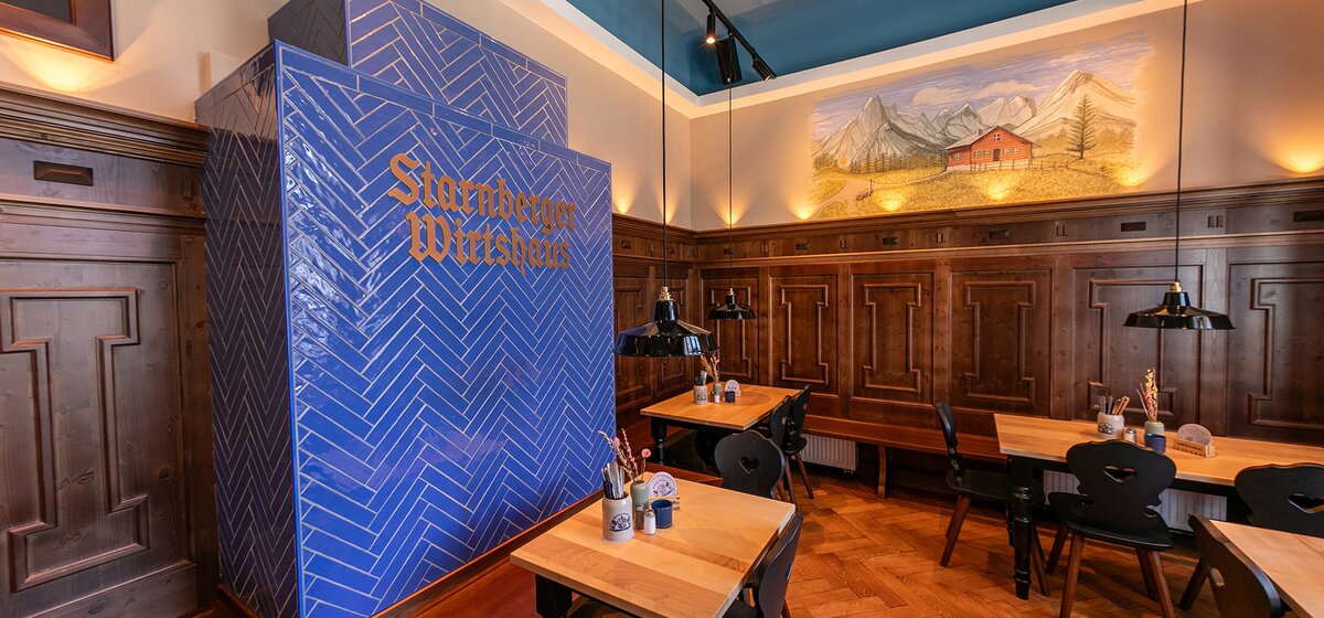 Innenraum des Starnberger Wirtshaus mit Holzwänden, Tischen und Stühlen, Wandbild mit Bergen und Haus. Im Zentrum der große blaue Kachelofen mit Starnberger Wirtshaus Schriftzug.