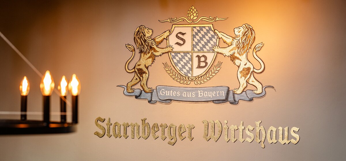 Wand mit Wappen und Schriftzug 'Starnberger Wirtshaus' und dem Motto 'Gutes aus Bayern', flankiert von zwei Löwen. Im Vordergrund ein unscharfer Kronleuchter. 