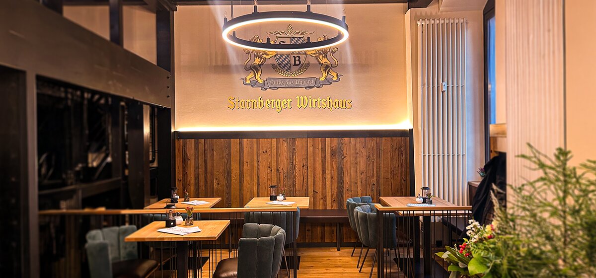 Wand mit Wappen und Schriftzug 'Starnberger Wirtshaus' und dem Motto 'Gutes aus Bayern'. Im Vordergrund die Gaststube mit Kronleuchter.